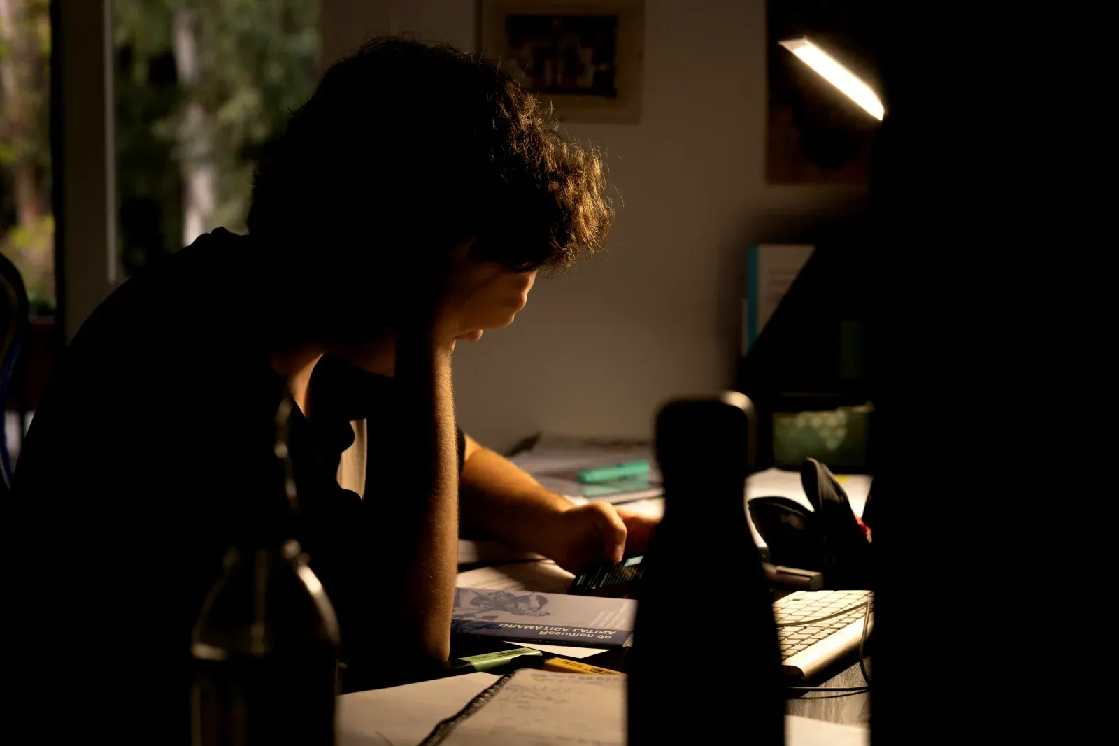 Personne assise de profil à un bureau sombre, éclairée par une lampe de table, la tête appuyée sur la main dans une attitude de fatigue ou de concentration intense.