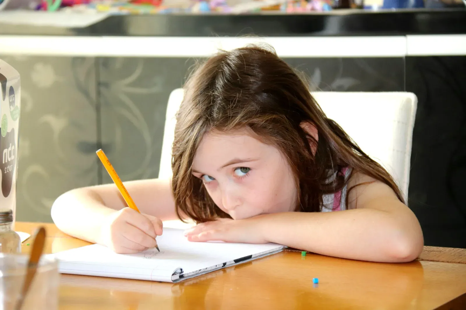 Une fillette aux cheveux longs bruns assise à une table en bois, écrivant ou dessinant dans un cahier avec un crayon jaune tout en regardant l'objectif avec lassitude.