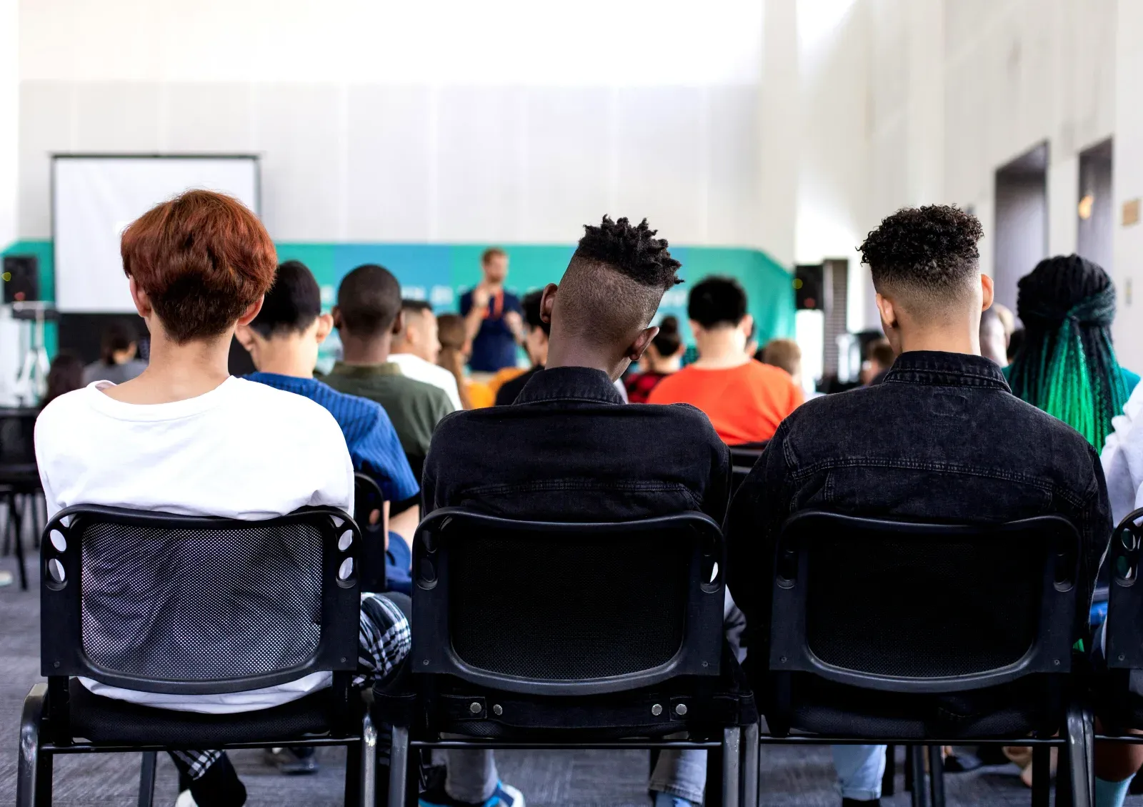 Élèves assis de dos sur des chaises dans une salle de classe, assistant à un cours ou une conférence face à un enseignant debout à l'avant.