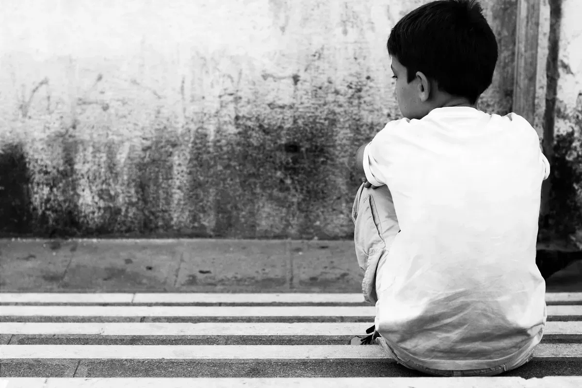 Photographie en noir et blanc d'un jeune garçon assis seul sur des marches en pierre, de dos, regardant le mur texturé et usé devant lui. Le garçon porte un t-shirt blanc froissé et un pantalon clair. L'ambiance est calme et contemplative, centrée sur sa silhouette isolée dans le coin droit.