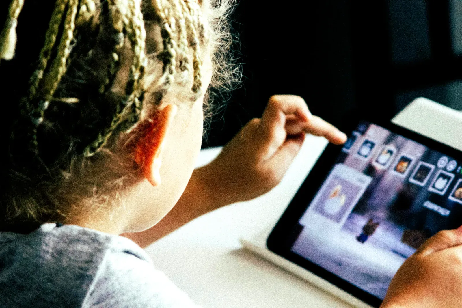 Un enfant aux cheveux tressés clairsemés de profil, interagissant avec l'écran tactile d'une tablette numérique affichant une application colorée.