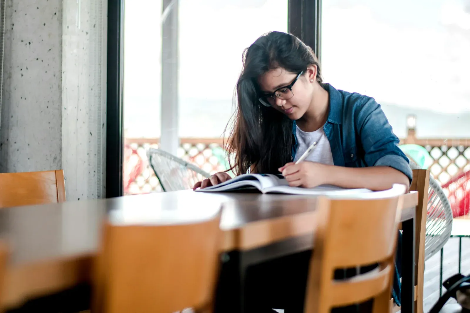 Jeune femme portant des lunettes et une chemise en jean, assise à une table en bois lumineuse, concentrée sur l'écriture dans un cahier ouvert.