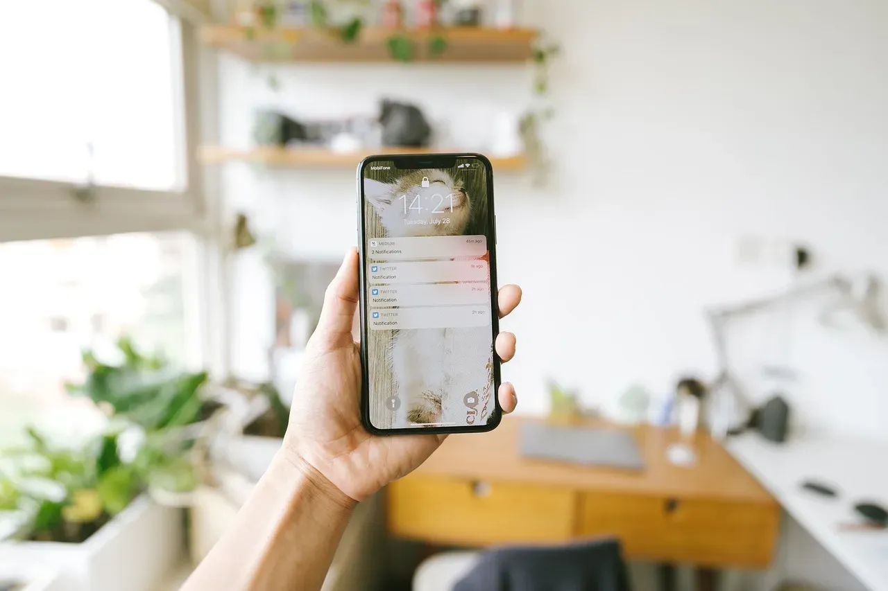Une main tient un smartphone dont l'écran affiche une application, devant un bureau et des étagères flous.