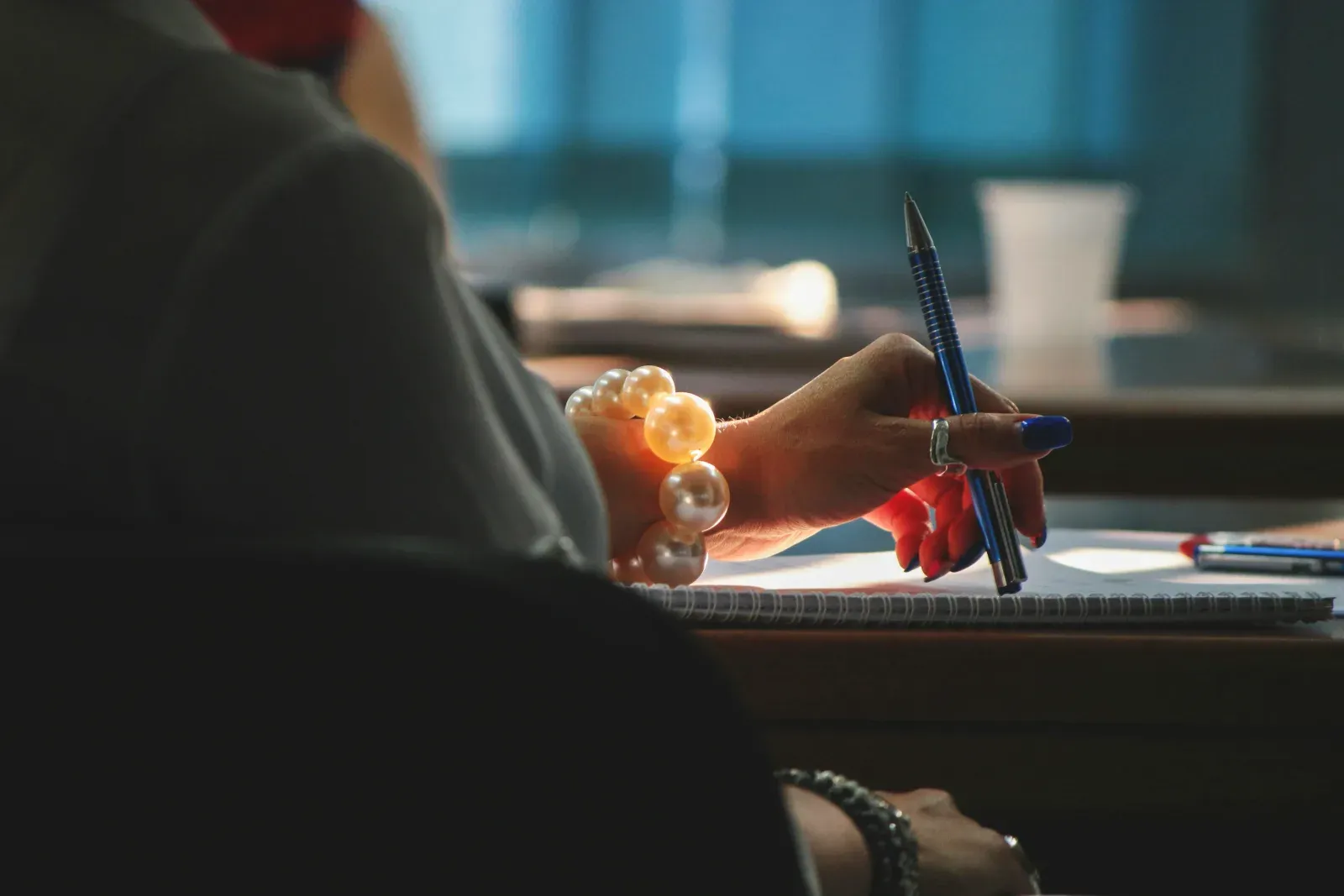 Gros plan sur la main d'une femme portant un bracelet de grosses perles claires, tenant un stylo bleu pour écrire sur un carnet posé sur un bureau.