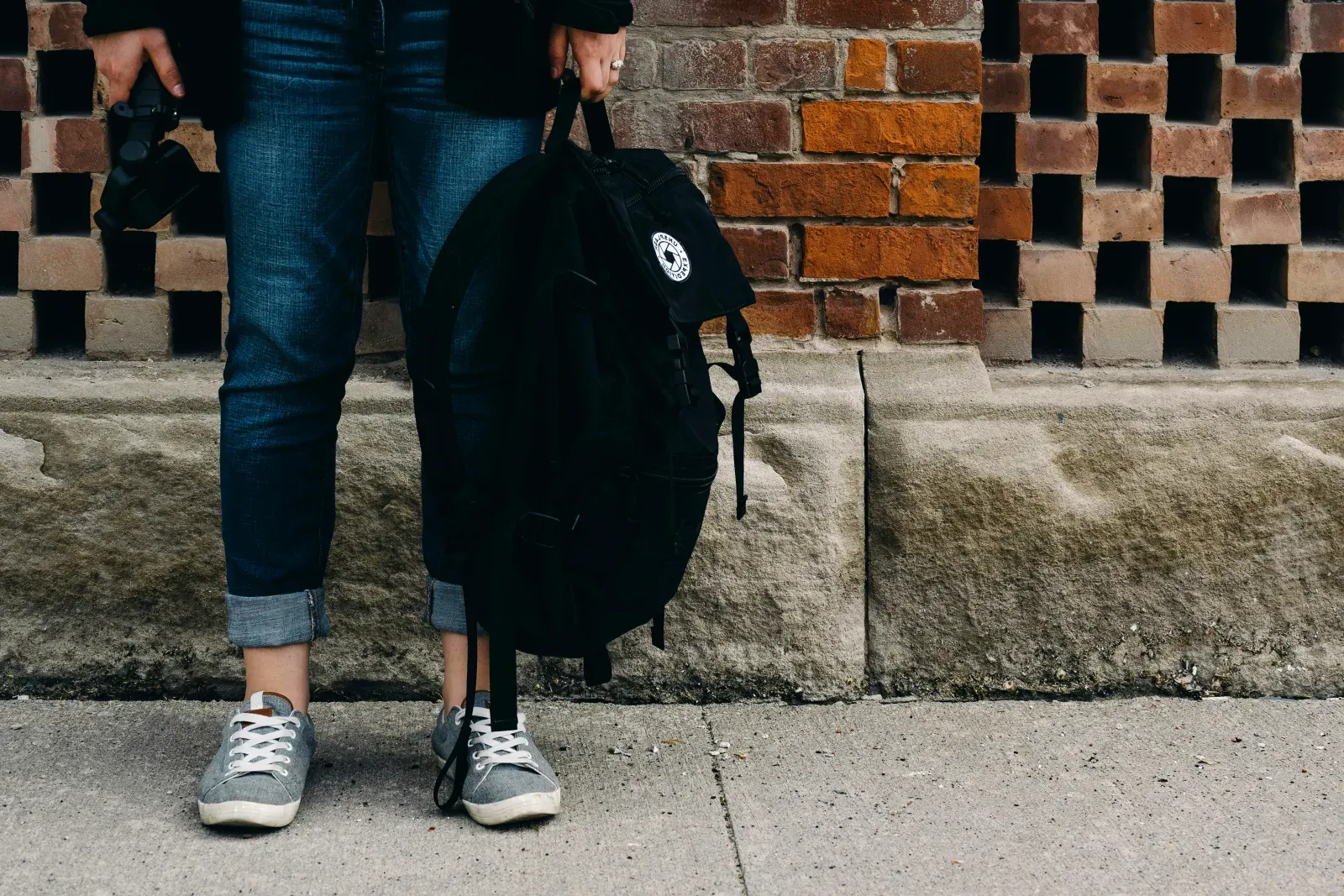 Vue de face, en plan serré sur les jambes et le bas du corps d'une personne en jean et baskets grises, tenant un sac à dos noir à la main devant un mur de briques ajouré.