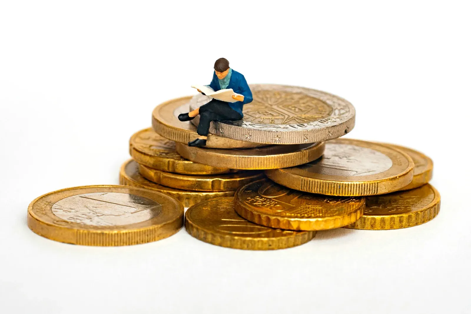 Figurine miniature d'une personne assise au sommet d'une pile de pièces de monnaie en euros, en train de lire un journal ouvert.